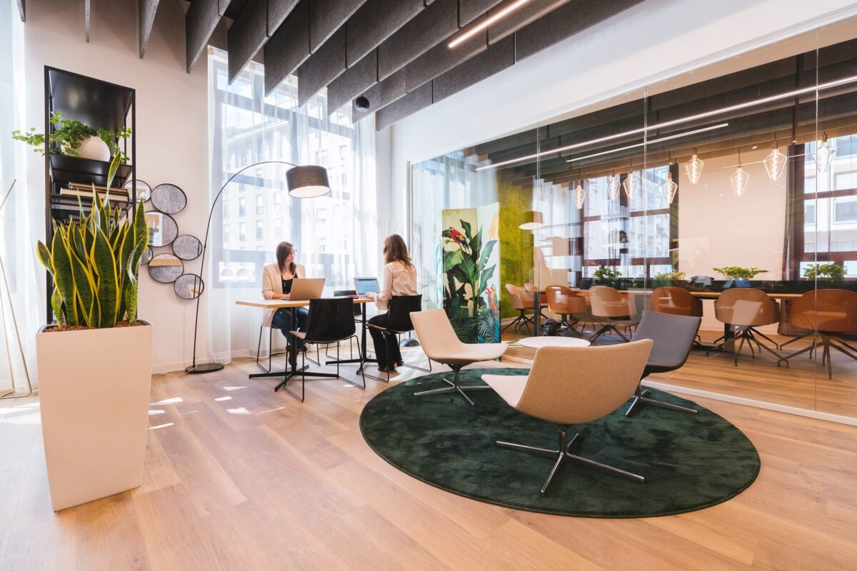 Un espace de bureau contemporain avec des panneaux acoustiques au plafond, de grandes fenêtres, des fauteuils lounge sur un tapis vert circulaire, des plantes d’intérieur et une salle de réunion aux parois vitrées où deux femmes travaillent sur des ordinateurs portables. L’espace est doté d’un parquet en bois clair et de meubles au design moderne.