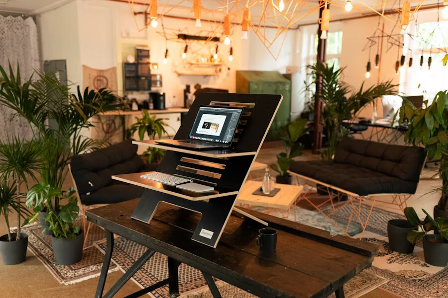 Coin de coworking cosy et éclectique avec bureau debout, fauteuils confortables, suspensions lumineuses et abondance de plantes en pot.